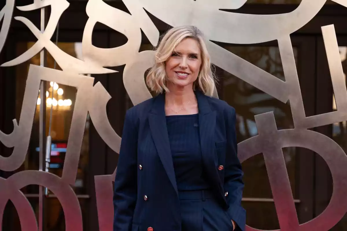 Una mujer rubia con traje oscuro sonríe frente a una escultura metálica abstracta. Una mujer rubia con traje oscuro sonríe frente a una escultura metálica abstracta.
