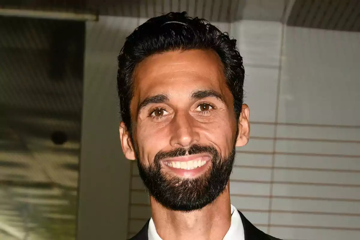 Un hombre con barba y cabello oscuro sonríe a la cámara en un entorno interior.