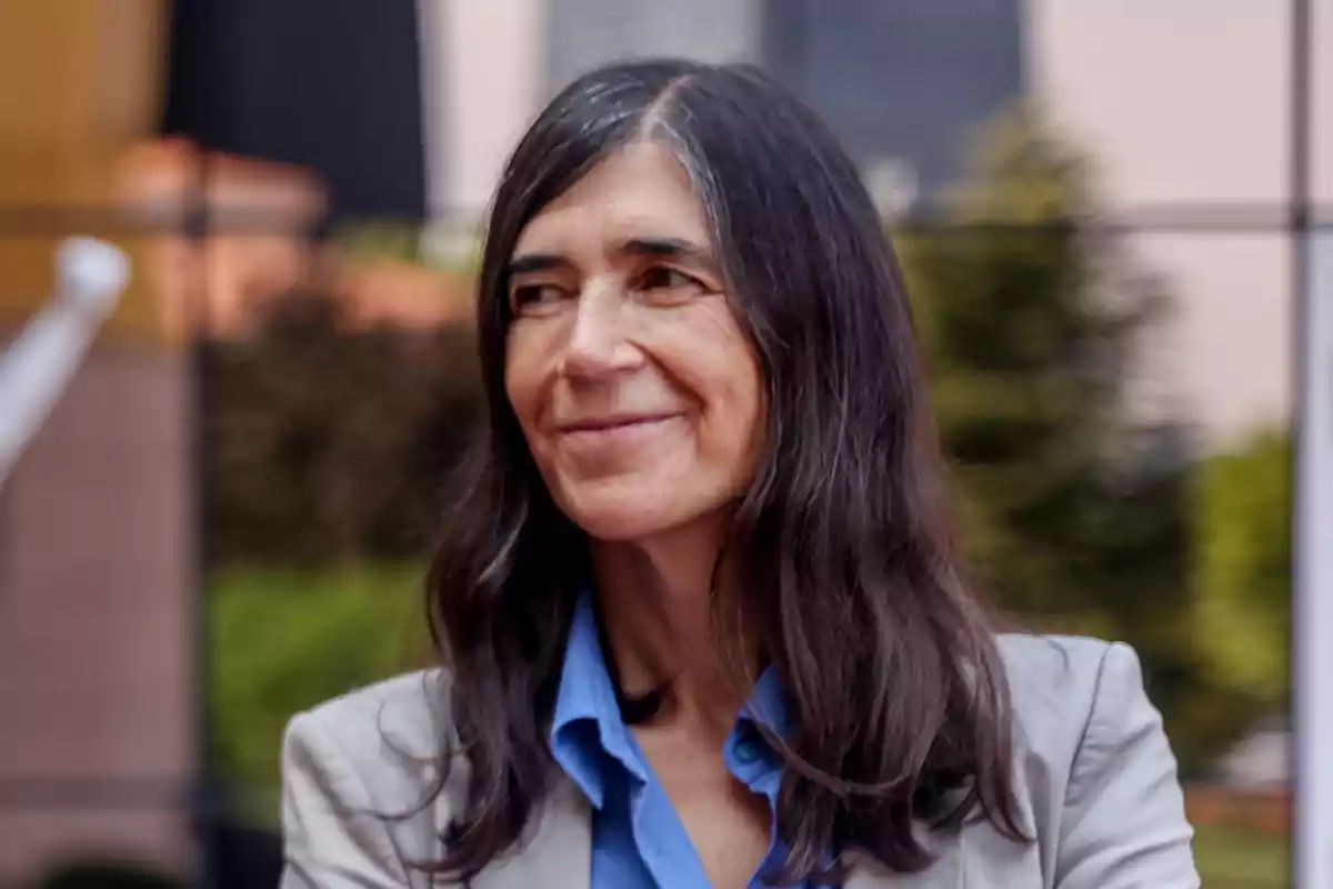 Una mujer de cabello largo y oscuro con una chaqueta clara y camisa azul sonriendo ligeramente al aire libre.