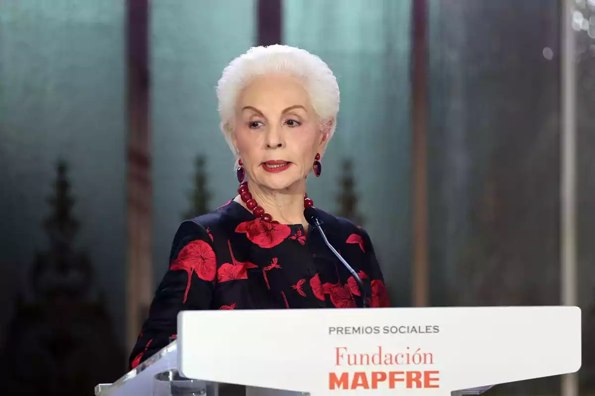 Una mujer mayor con cabello blanco y un collar rojo habla en un podio durante un evento de la Fundación Mapfre.