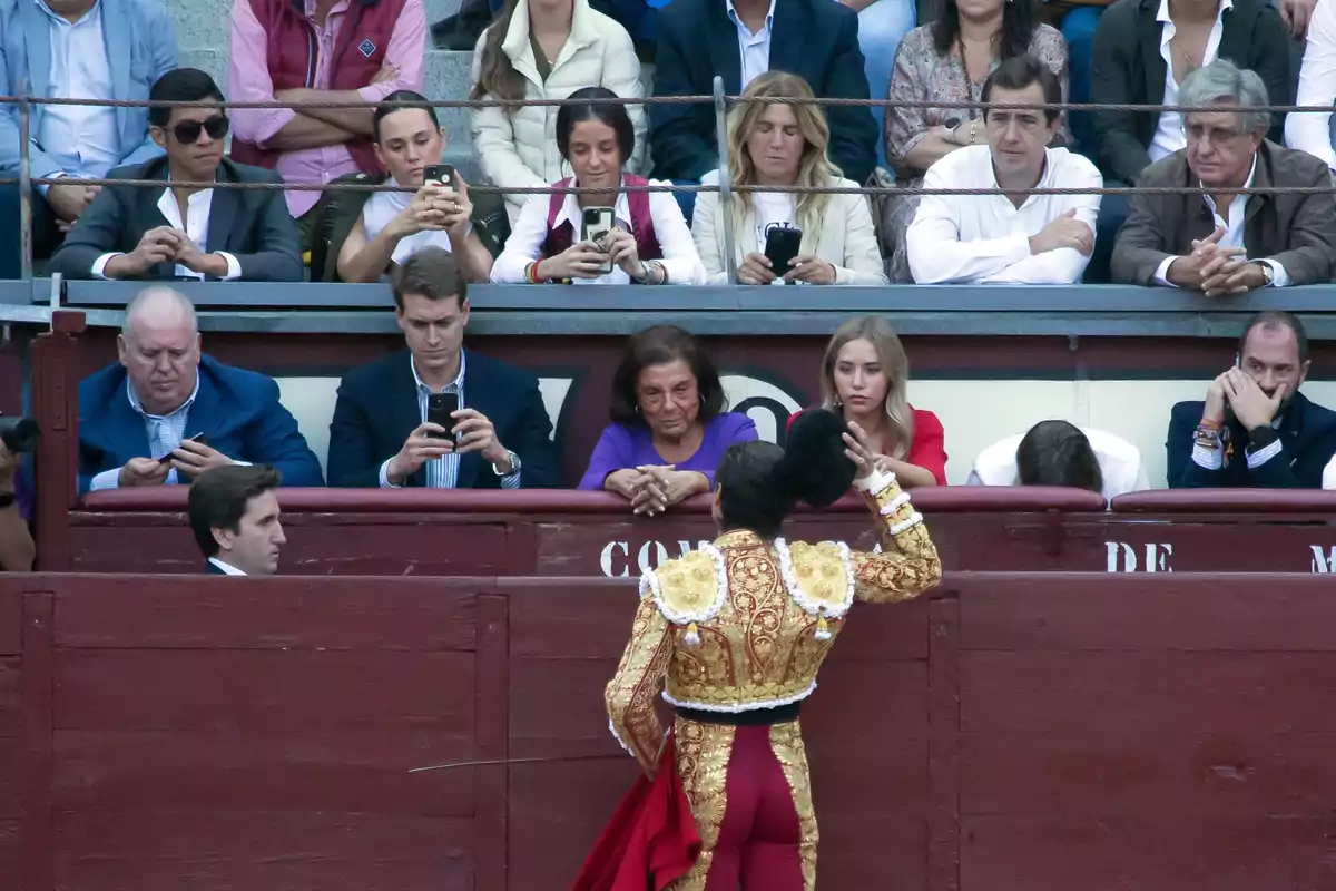 Un torero saluda a los espectadores en una plaza de toros, mientras varias personas en las gradas observan y usan sus teléfonos móviles.