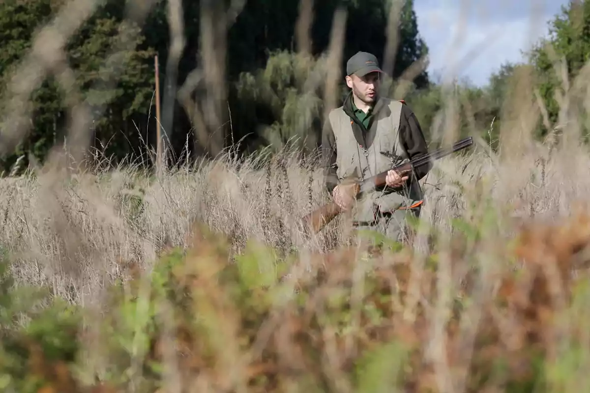 Hombre con ropa de caza y gorra sosteniendo una escopeta en un campo con vegetación seca y árboles al fondo.