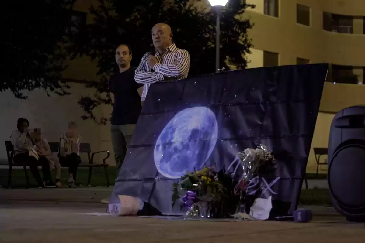 Dos personas de pie junto a una imagen de la luna y flores en un espacio al aire libre por la noche.