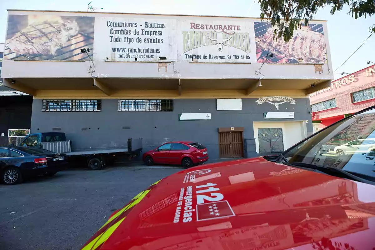 Fachada de un restaurante llamado Rancho Rodeo con un gran cartel publicitario y varios coches aparcados frente al edificio, incluyendo un vehículo de emergencias de la Comunidad de Madrid en primer plano Fachada de un restaurante llamado Rancho Rodeo con un gran cartel publicitario y varios coches aparcados frente al edificio, incluyendo un vehículo de emergencias de la Comunidad de Madrid en primer plano