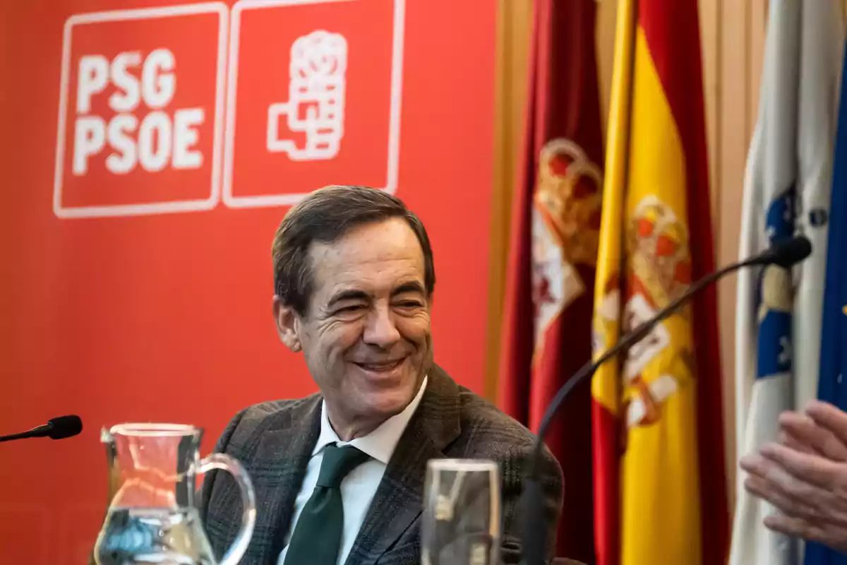 Un hombre sonríe mientras está sentado frente a un fondo rojo con el logotipo del PSG PSOE y varias banderas detrás. Un hombre sonríe mientras está sentado frente a un fondo rojo con el logotipo del PSG PSOE y varias banderas detrás.
