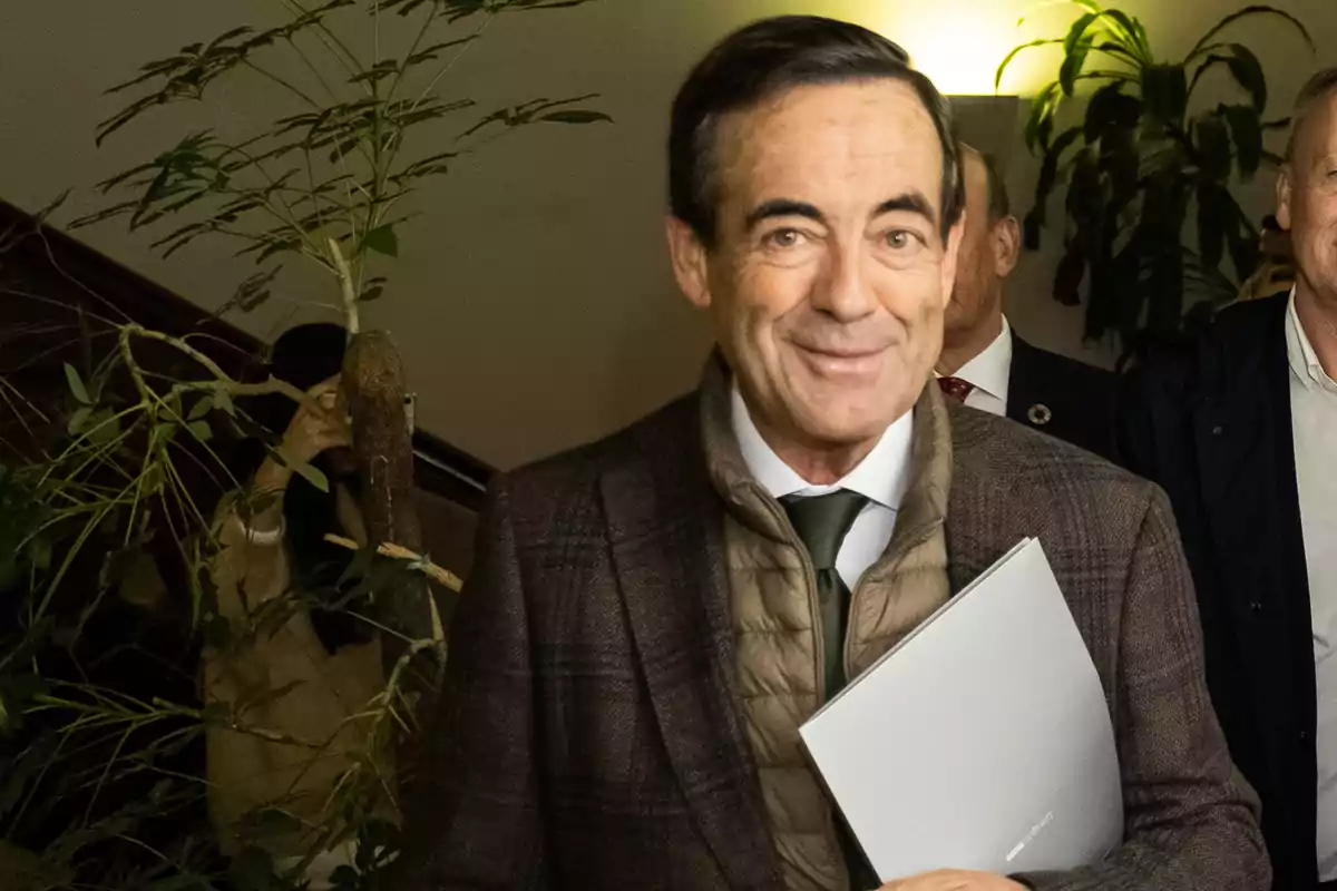 Un hombre sonriente con chaqueta marrón y corbata verde sostiene un documento mientras camina por un pasillo con plantas al fondo. Un hombre sonriente con chaqueta marrón y corbata verde sostiene un documento mientras camina por un pasillo con plantas al fondo.