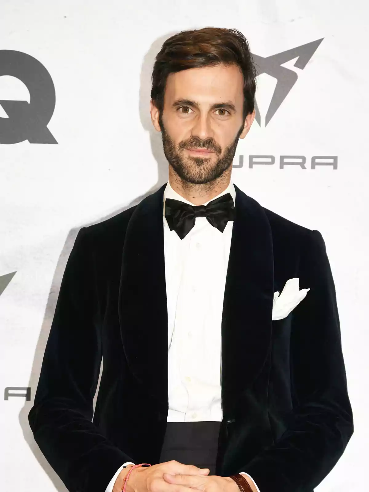 Un hombre con esmoquin negro y pajarita posa frente a un fondo con logotipos. Un hombre con esmoquin negro y pajarita posa frente a un fondo con logotipos.