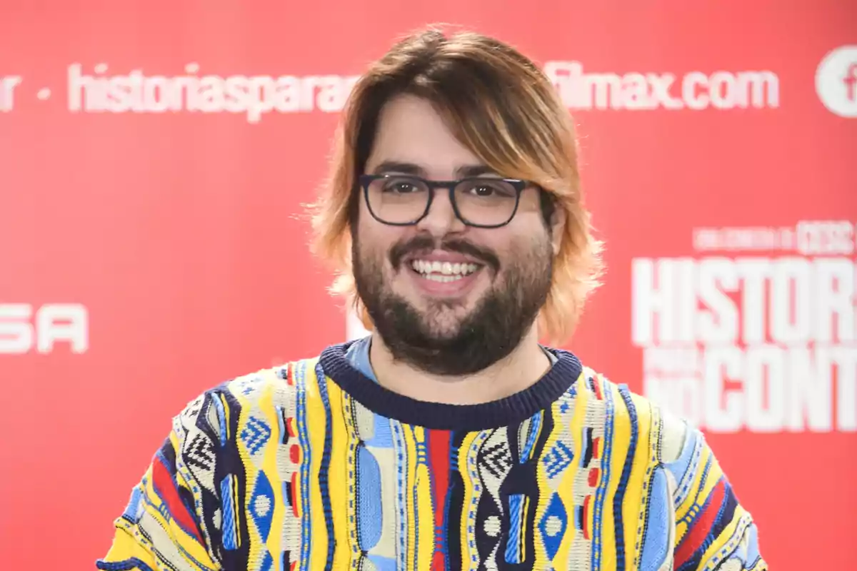 Un hombre con gafas y barba sonríe frente a un fondo rojo con texto blanco, vistiendo un suéter colorido con patrones geométricos.