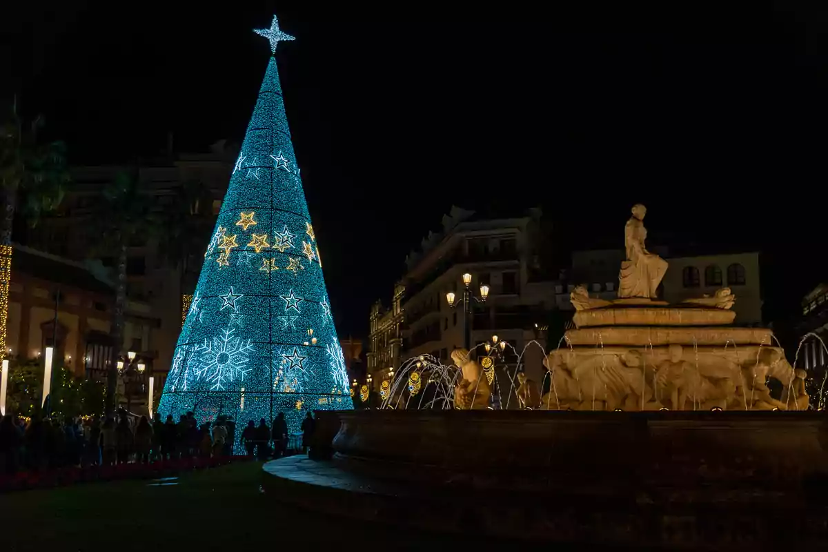 Un árbol de Navidad iluminado con estrellas y copos de nieve junto a una fuente en una plaza por la noche.