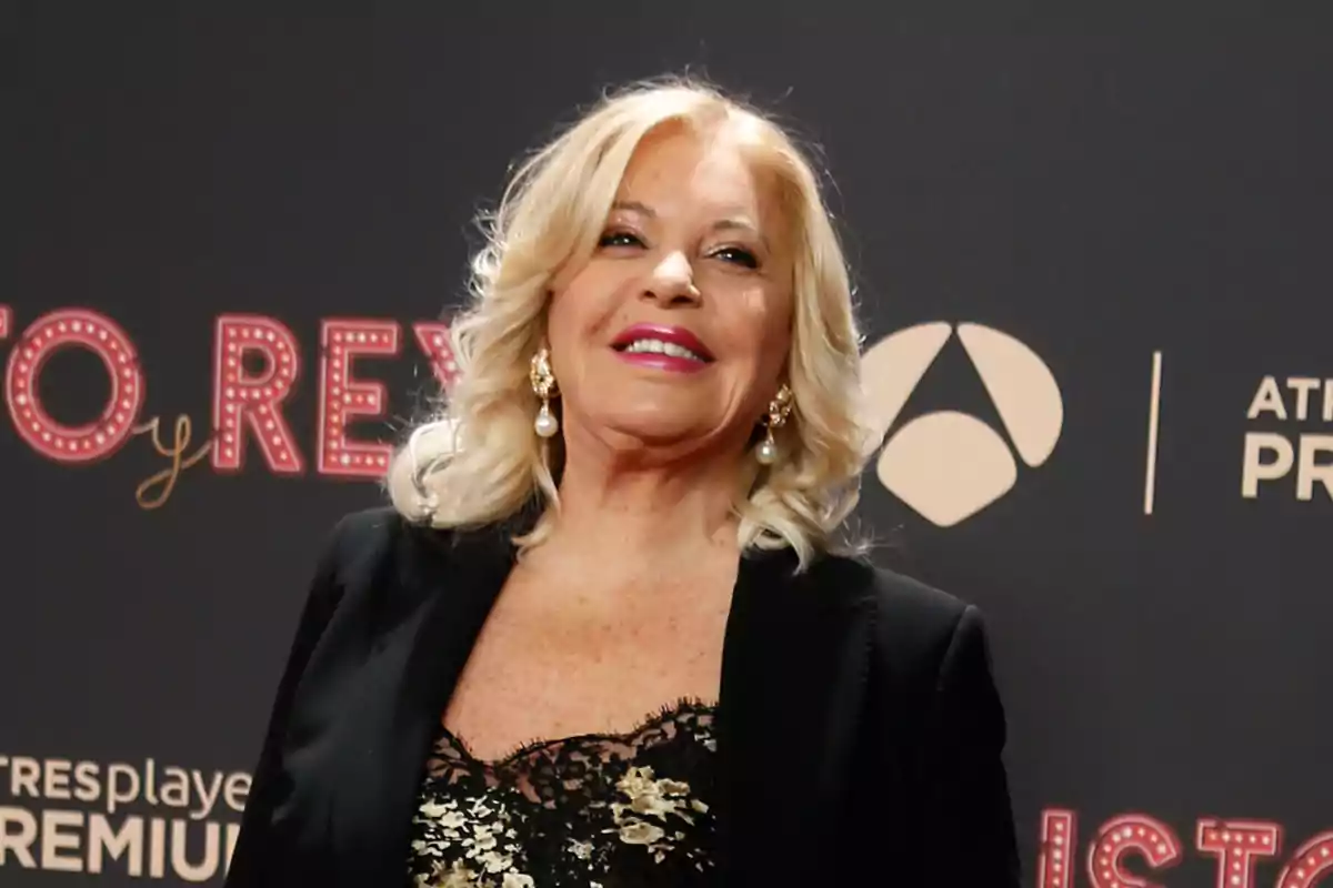 Una mujer rubia sonriente con un vestido negro y detalles dorados en un evento con fondo de logotipos.