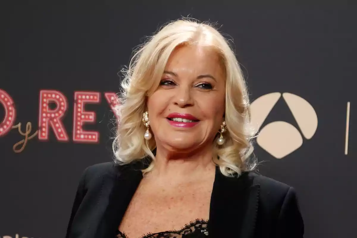 Una mujer rubia con cabello rizado y maquillaje sonríe mientras lleva un vestido negro elegante y pendientes de perlas en un evento con un fondo oscuro.