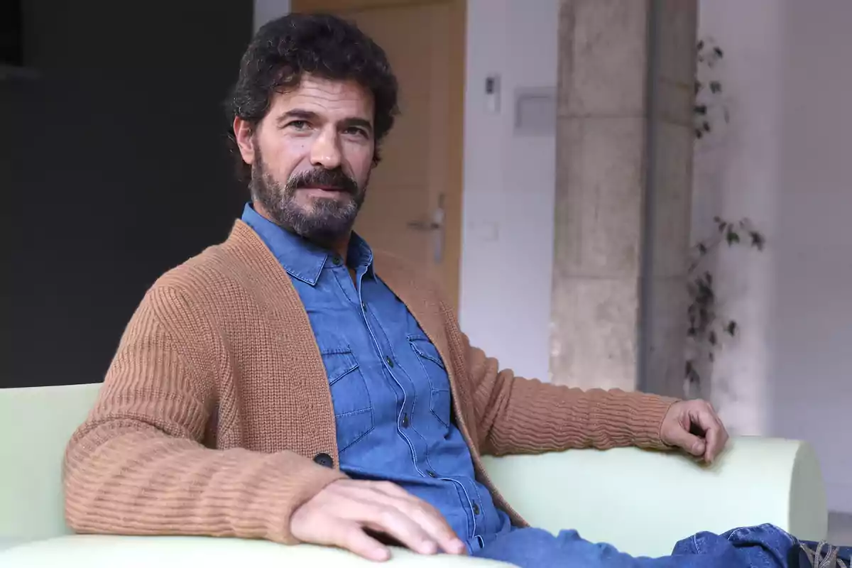 Un hombre con barba y cabello oscuro está sentado en un sofá verde, vistiendo una camisa de mezclilla y un cárdigan marrón, en un ambiente interior con paredes claras y una planta al fondo.