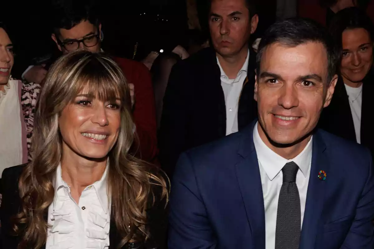 Una mujer y un hombre sonriendo en un evento con más personas en el fondo.