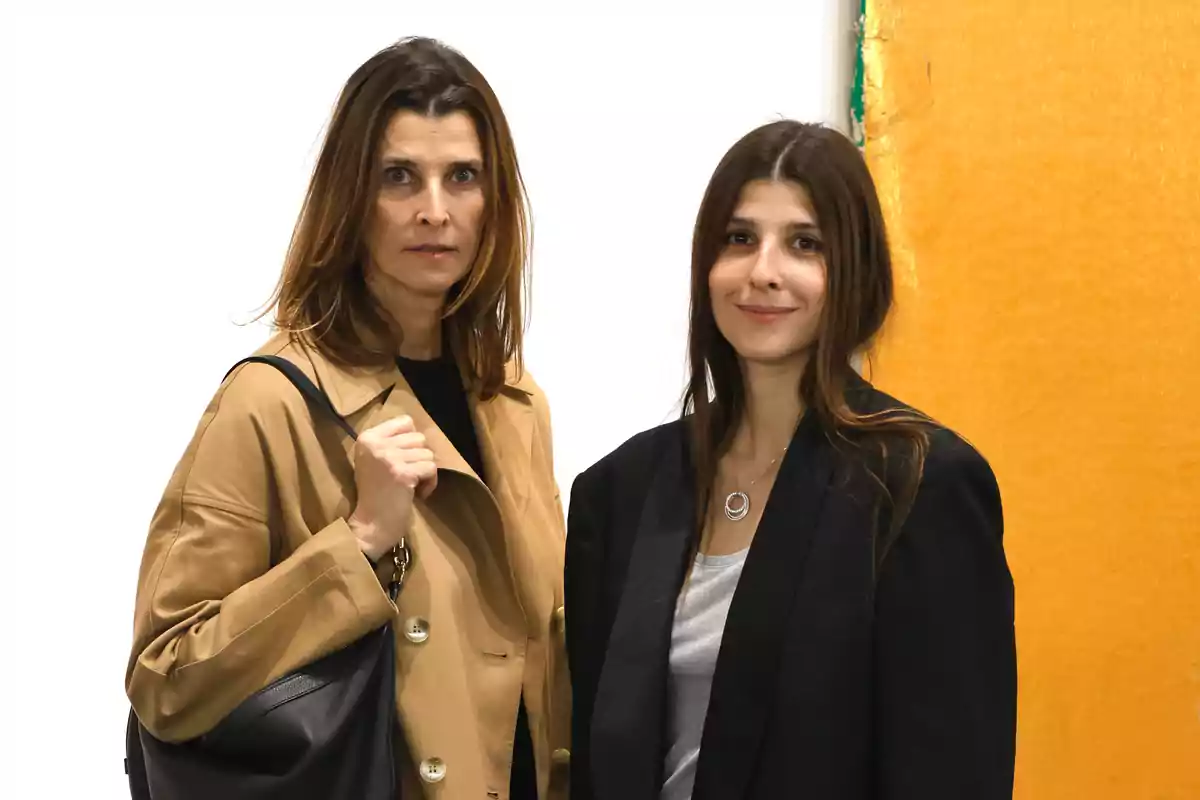 Dos mujeres posan juntas frente a un fondo blanco y amarillo, una lleva un abrigo beige y la otra un blazer negro.