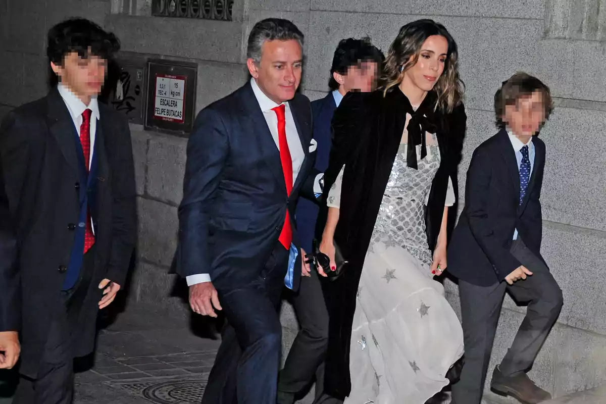 Un grupo de personas vestidas formalmente camina por una acera junto a un edificio de piedra. Un grupo de personas vestidas formalmente camina por una acera junto a un edificio de piedra.