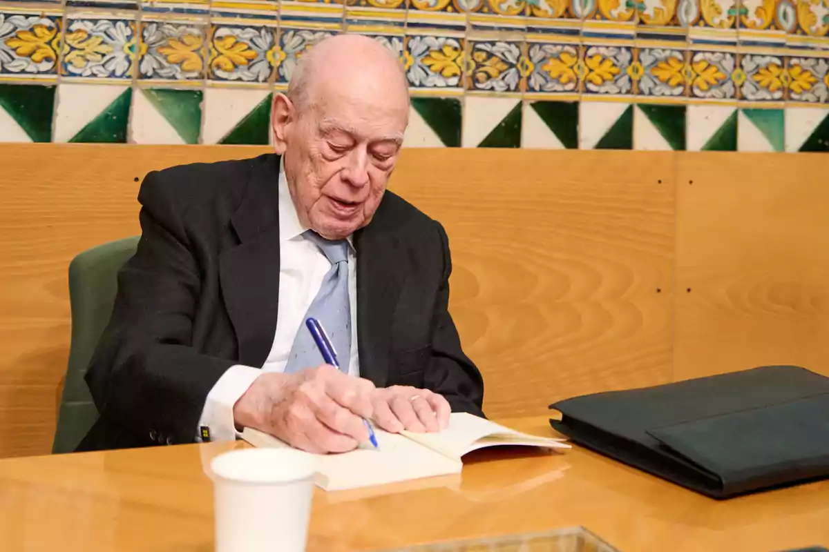 Un hombre mayor firmando un libro en una mesa de madera con un fondo de azulejos decorativos.