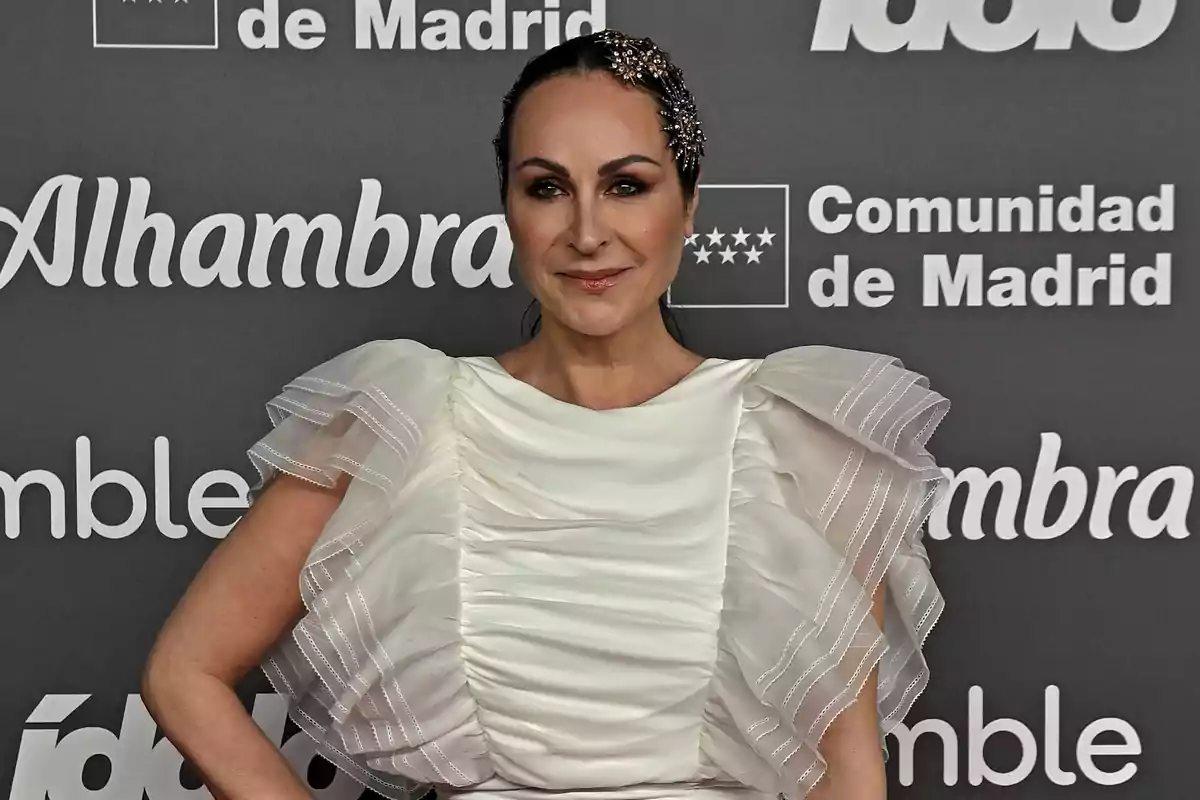 Una persona con un vestido blanco elegante posando frente a un fondo con logotipos de Alhambra y Comunidad de Madrid.