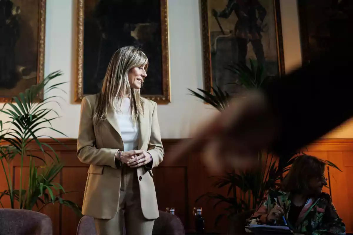 Una mujer de pie con traje beige y blusa blanca, sonriendo y mirando hacia la derecha, mientras otra persona está sentada en el fondo tomando notas, en una sala decorada con plantas y cuadros en la pared.