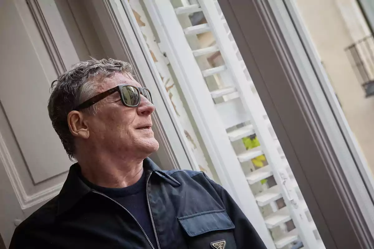 Hombre de cabello canoso y gafas de sol mirando por una ventana con persianas blancas, vestido con chaqueta oscura Hombre de cabello canoso y gafas de sol mirando por una ventana con persianas blancas, vestido con chaqueta oscura
