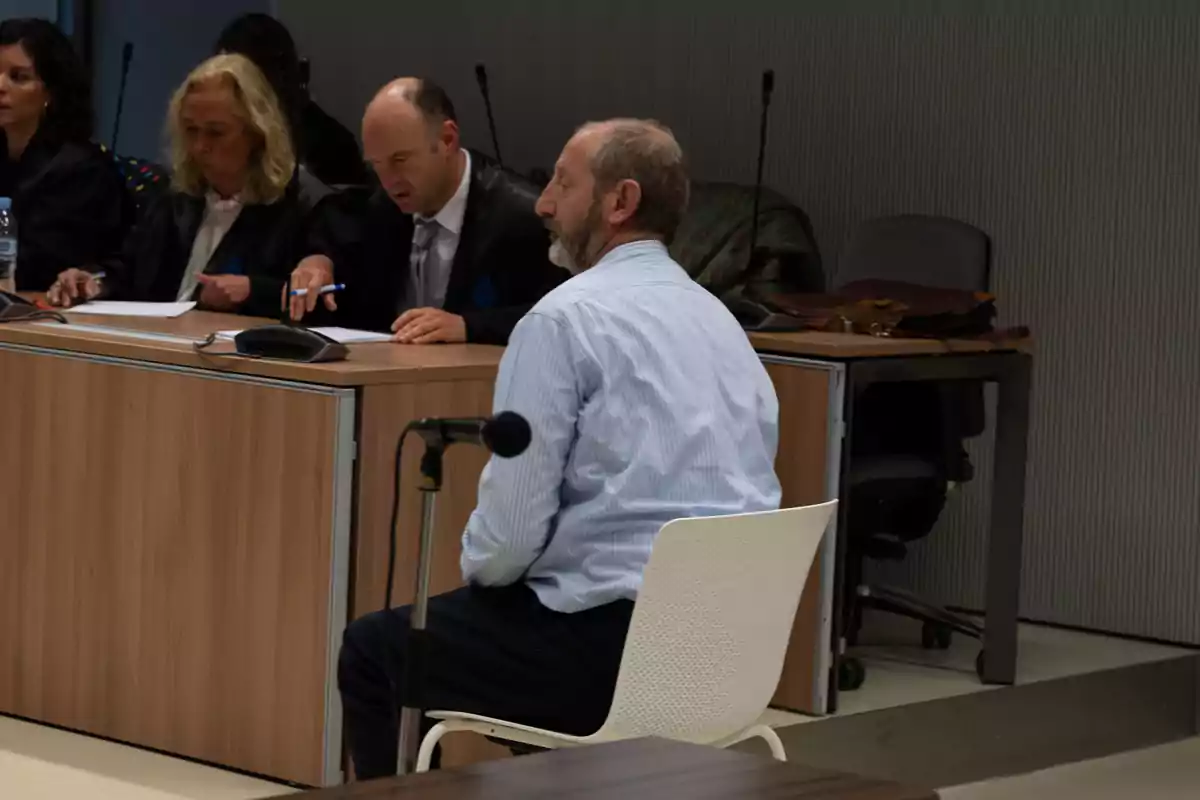 Un hombre sentado frente a un tribunal con tres personas revisando documentos en una sala de audiencias.