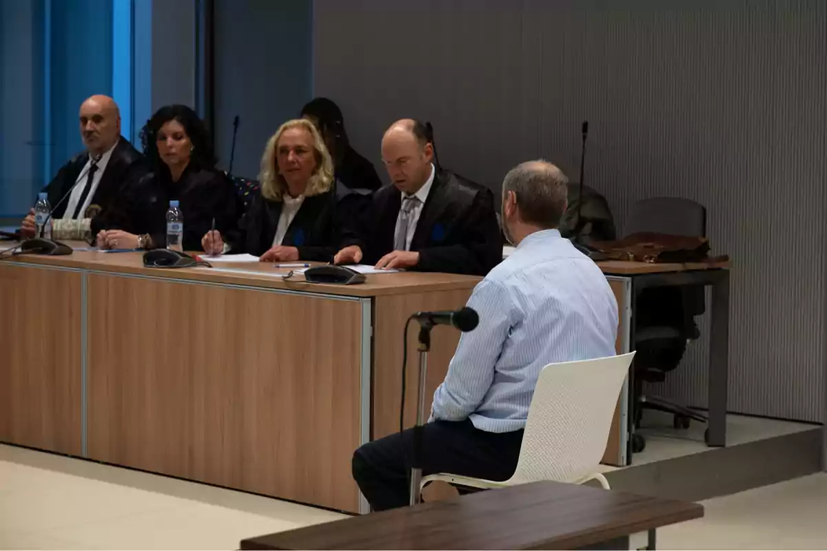 Un hombre sentado frente a un panel de jueces en una sala de audiencias. Un hombre sentado frente a un panel de jueces en una sala de audiencias.