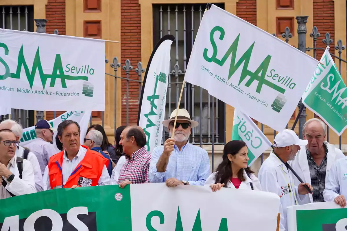 Grupo de personas manifestándose con batas blancas y banderas del Sindicato Médico Andaluz frente a un edificio antiguo Grupo de personas manifestándose con batas blancas y banderas del Sindicato Médico Andaluz frente a un edificio antiguo