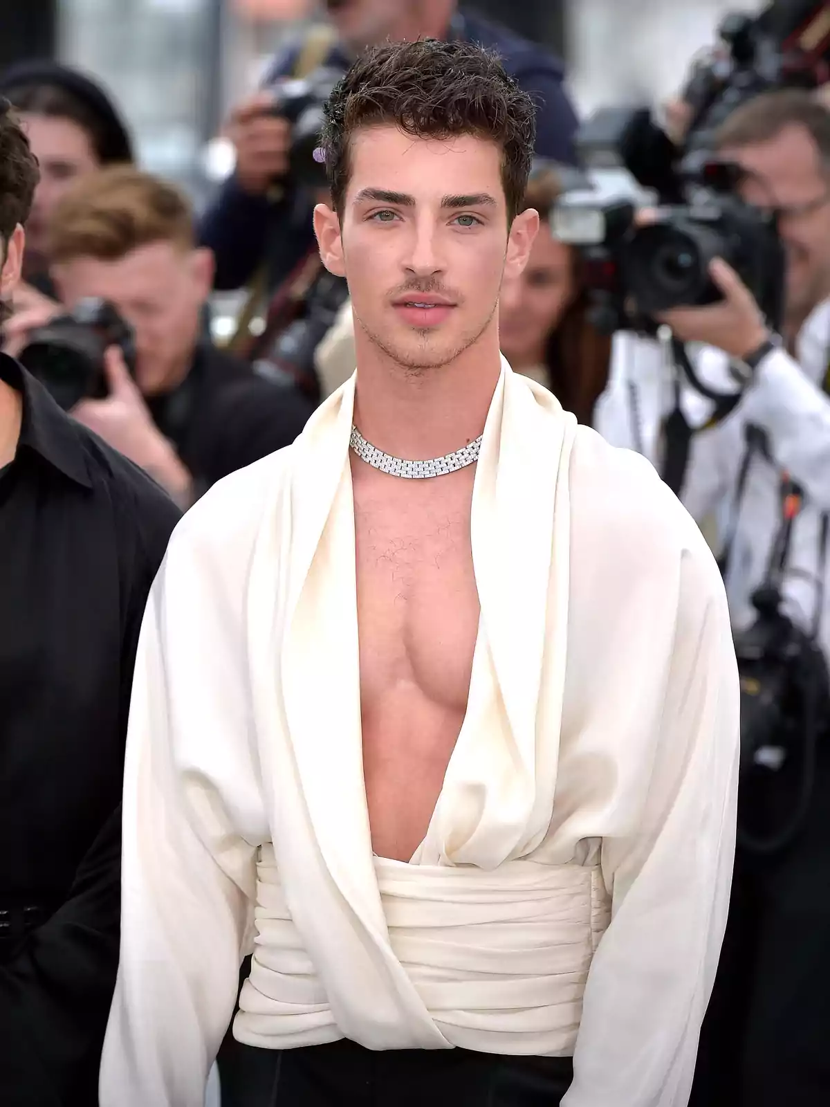 Un hombre con una camisa blanca de estilo único y un collar de diamantes, posando frente a varias cámaras en un evento.