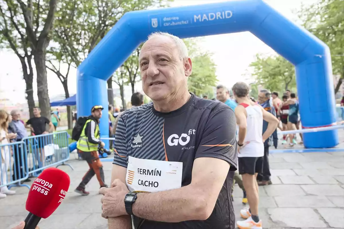 Un hombre con camiseta deportiva y un dorsal en el pecho está de pie frente a un arco inflable azul en un evento al aire libre, mientras una persona sostiene un micrófono rojo cerca de él.