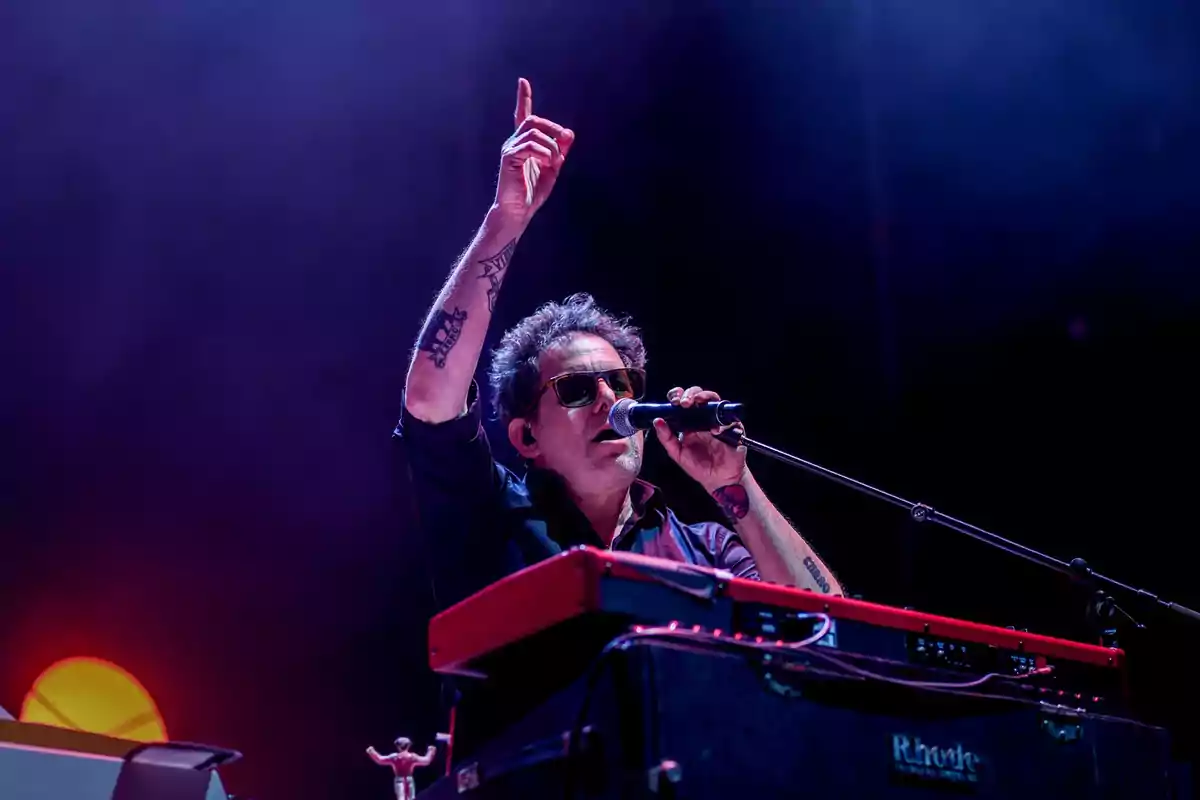Un músico con gafas de sol canta y toca el teclado en un escenario iluminado.