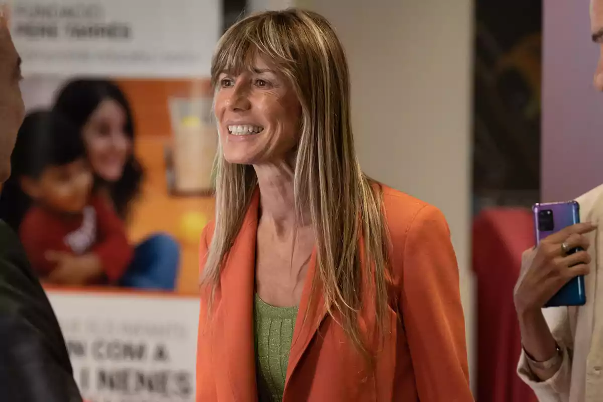 Una mujer sonriente con cabello largo y rubio lleva un blazer naranja y una camiseta verde mientras conversa con otras personas en un evento.