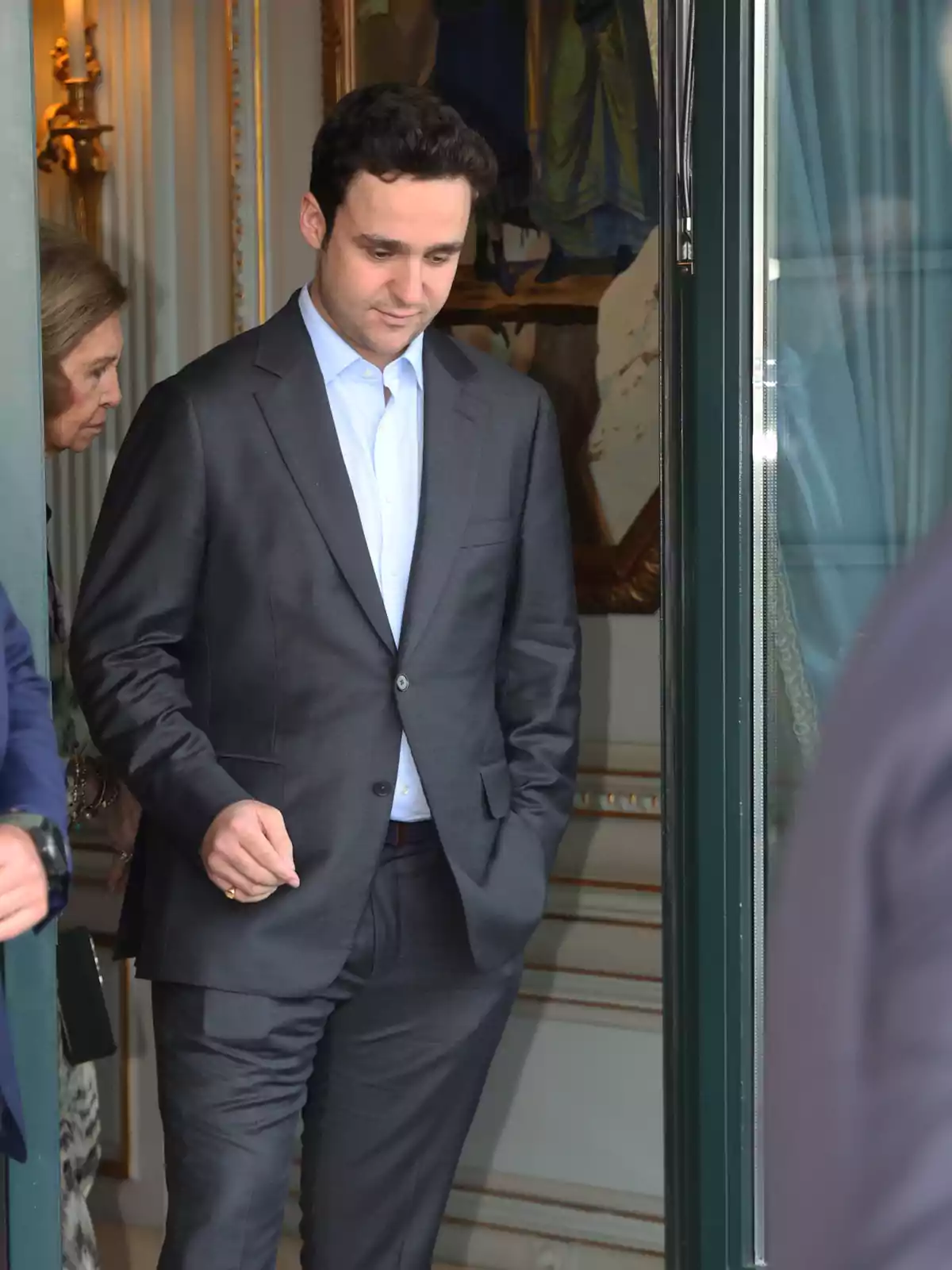Hombre joven con traje gris y camisa azul claro saliendo por una puerta de cristal mientras una mujer mayor lo sigue desde atrás en un entorno elegante Hombre joven con traje gris y camisa azul claro saliendo por una puerta de cristal mientras una mujer mayor lo sigue desde atrás en un entorno elegante