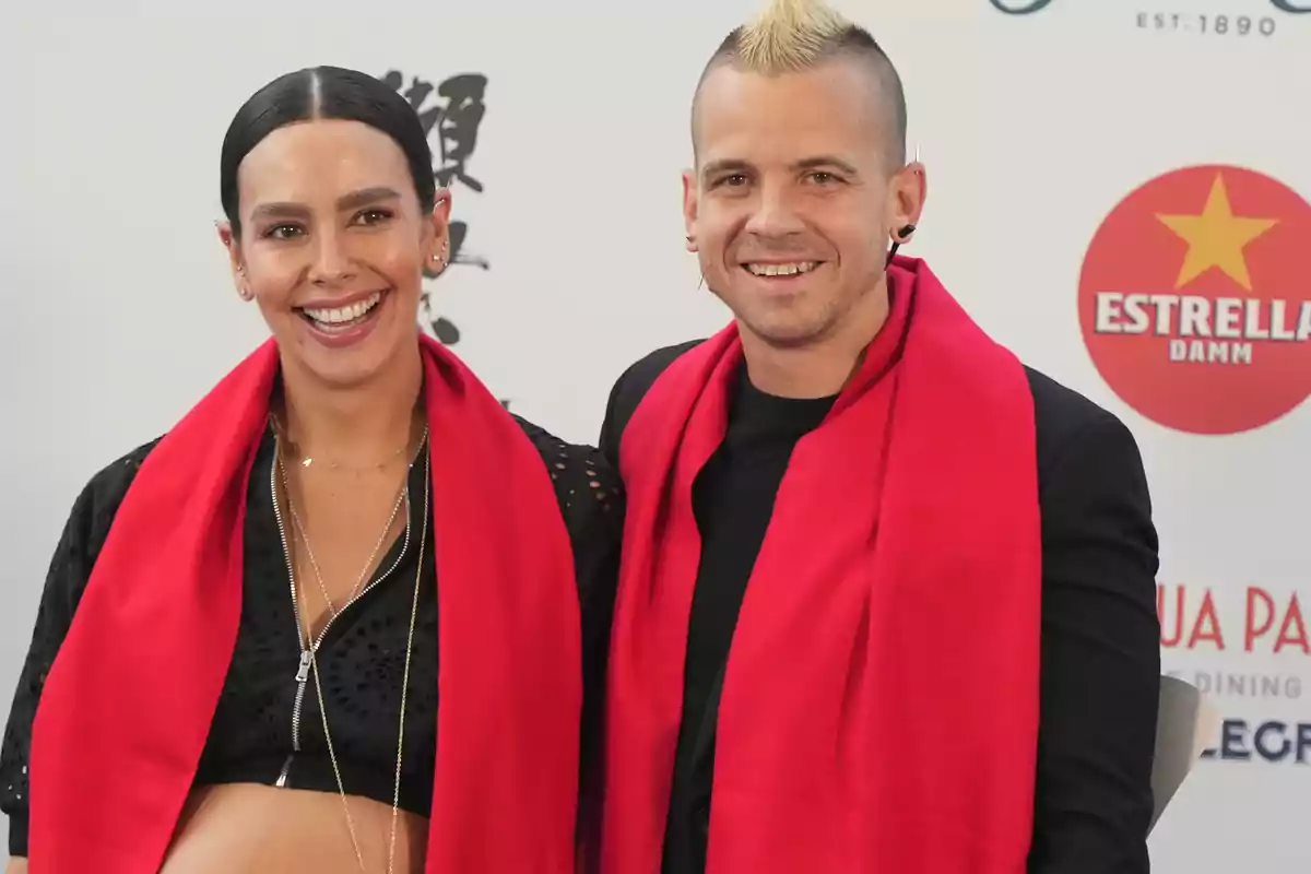 Una pareja sonriente con bufandas rojas posando frente a un fondo con logotipos.