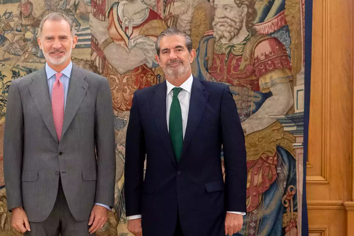 Dos hombres posan juntos frente a un tapiz decorativo.