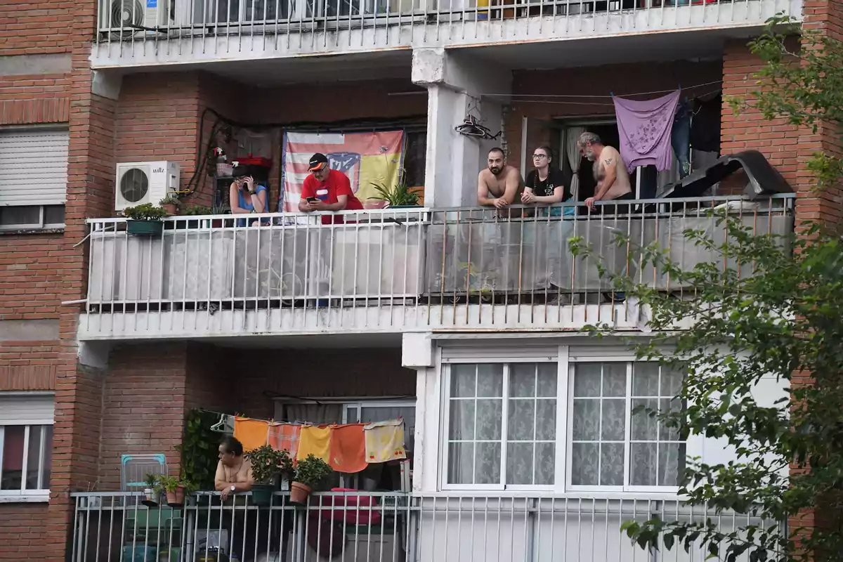 Varias personas están en los balcones de un edificio de ladrillo, algunas mirando hacia afuera y otras usando dispositivos electrónicos, con ropa colgada y plantas en las barandillas.