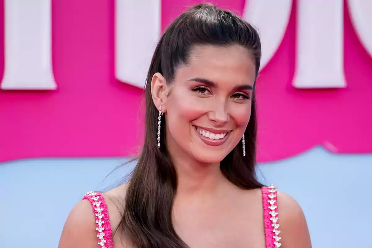 Una mujer con cabello largo y oscuro, usando un vestido rosa brillante y pendientes largos, sonríe frente a un fondo rosa y azul.