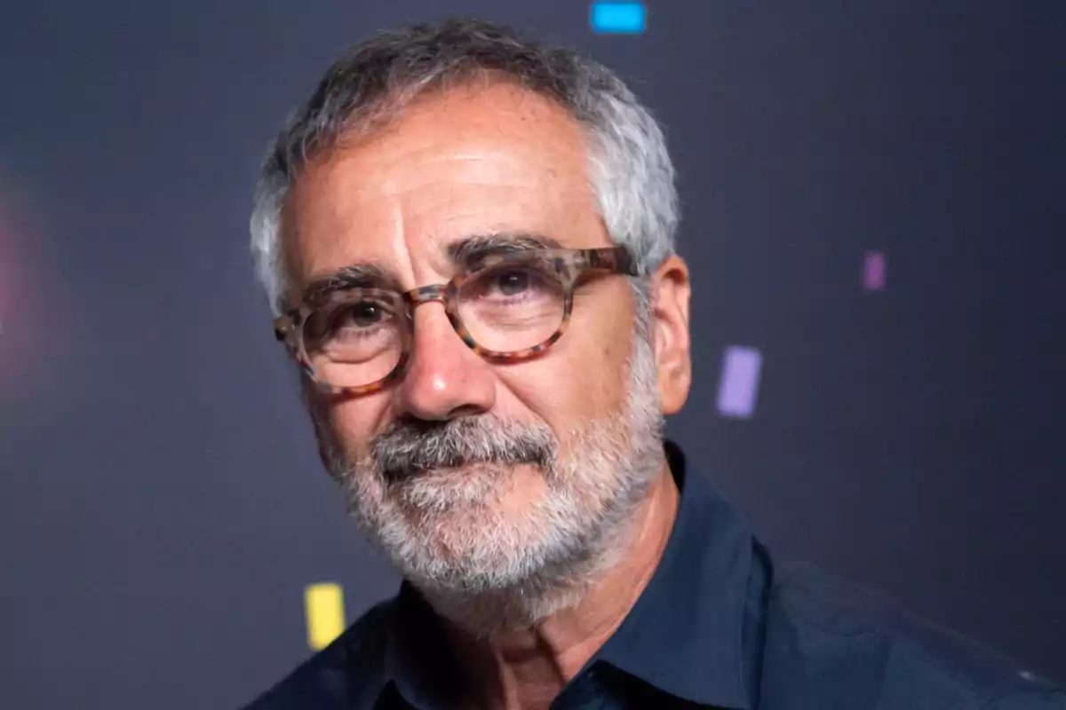 Un hombre con barba y gafas de montura gruesa frente a un fondo oscuro con formas de colores.