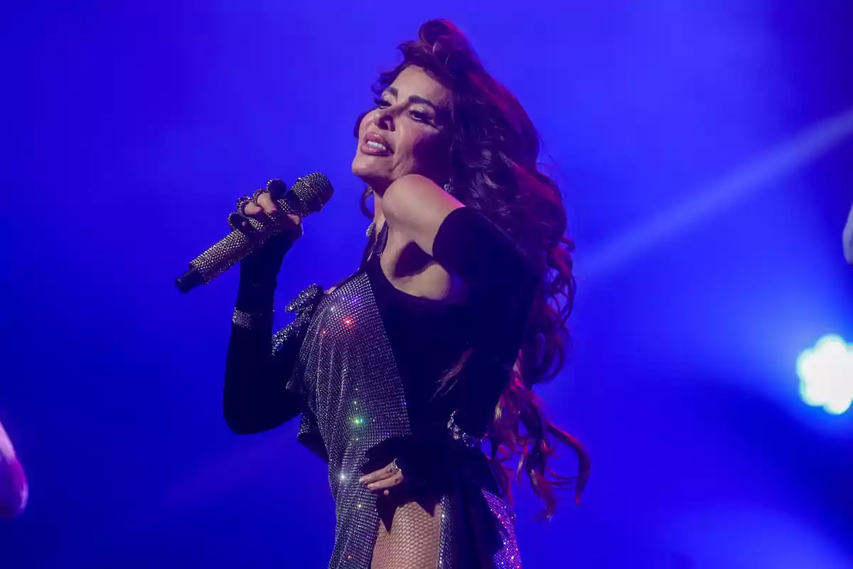 Cantante con vestido brillante y guantes negros sosteniendo un micrófono durante una presentación en el escenario con luces azules de fondo.