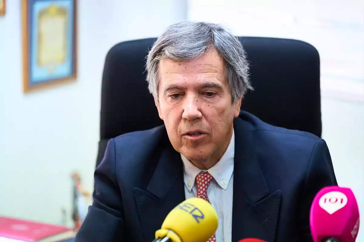 Hombre de cabello canoso y traje oscuro sentado en una oficina frente a varios micrófonos de prensa Hombre de cabello canoso y traje oscuro sentado en una oficina frente a varios micrófonos de prensa