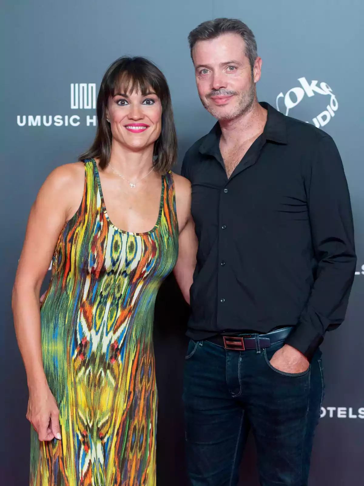 Una mujer con un vestido colorido y un hombre con camisa negra posan juntos frente a un fondo con logotipos.