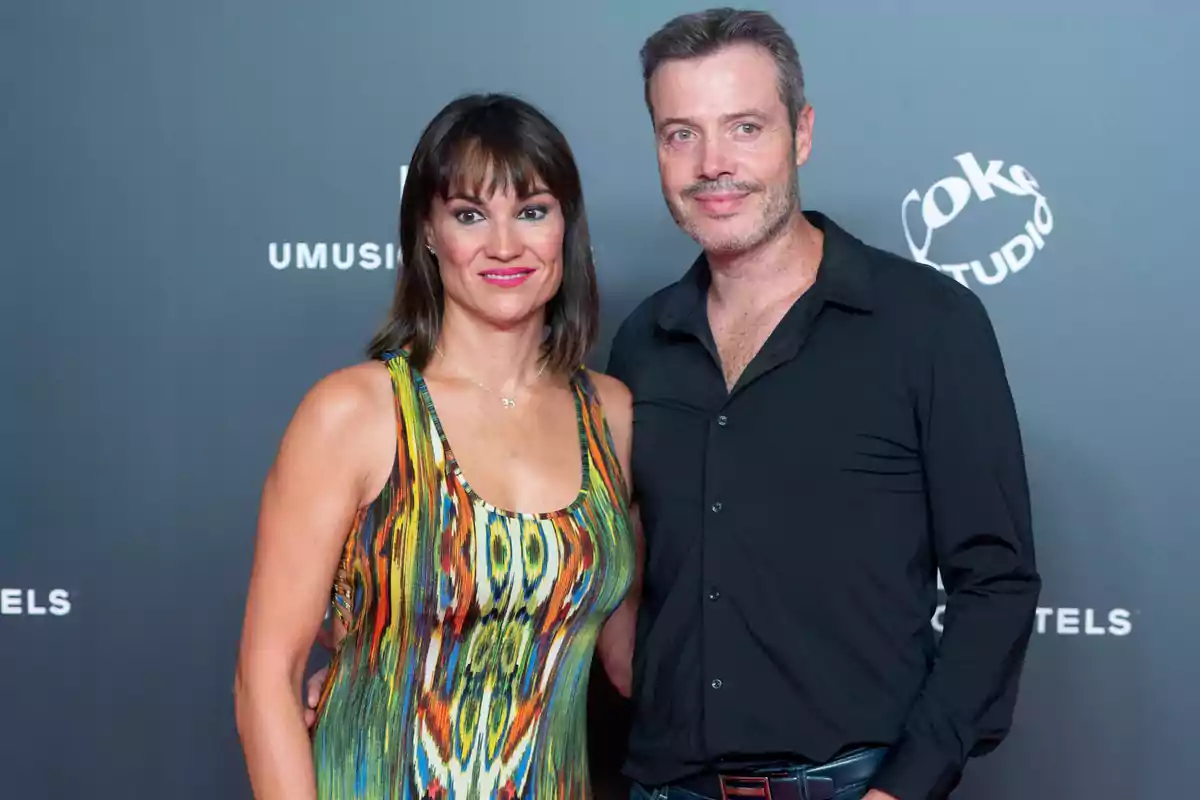 Una mujer con un vestido colorido y un hombre con camisa negra posan juntos frente a un fondo gris con logotipos.