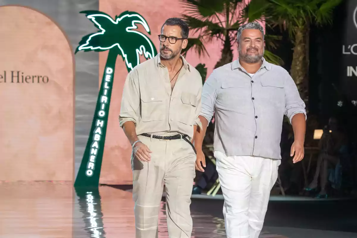 Dos hombres caminando juntos en una pasarela de moda con un fondo de palmeras y un letrero que dice 