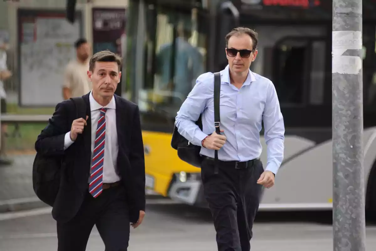 Dos hombres caminando por la calle, uno con traje y corbata y el otro con camisa azul y gafas de sol, con un autobús amarillo y negro de fondo.