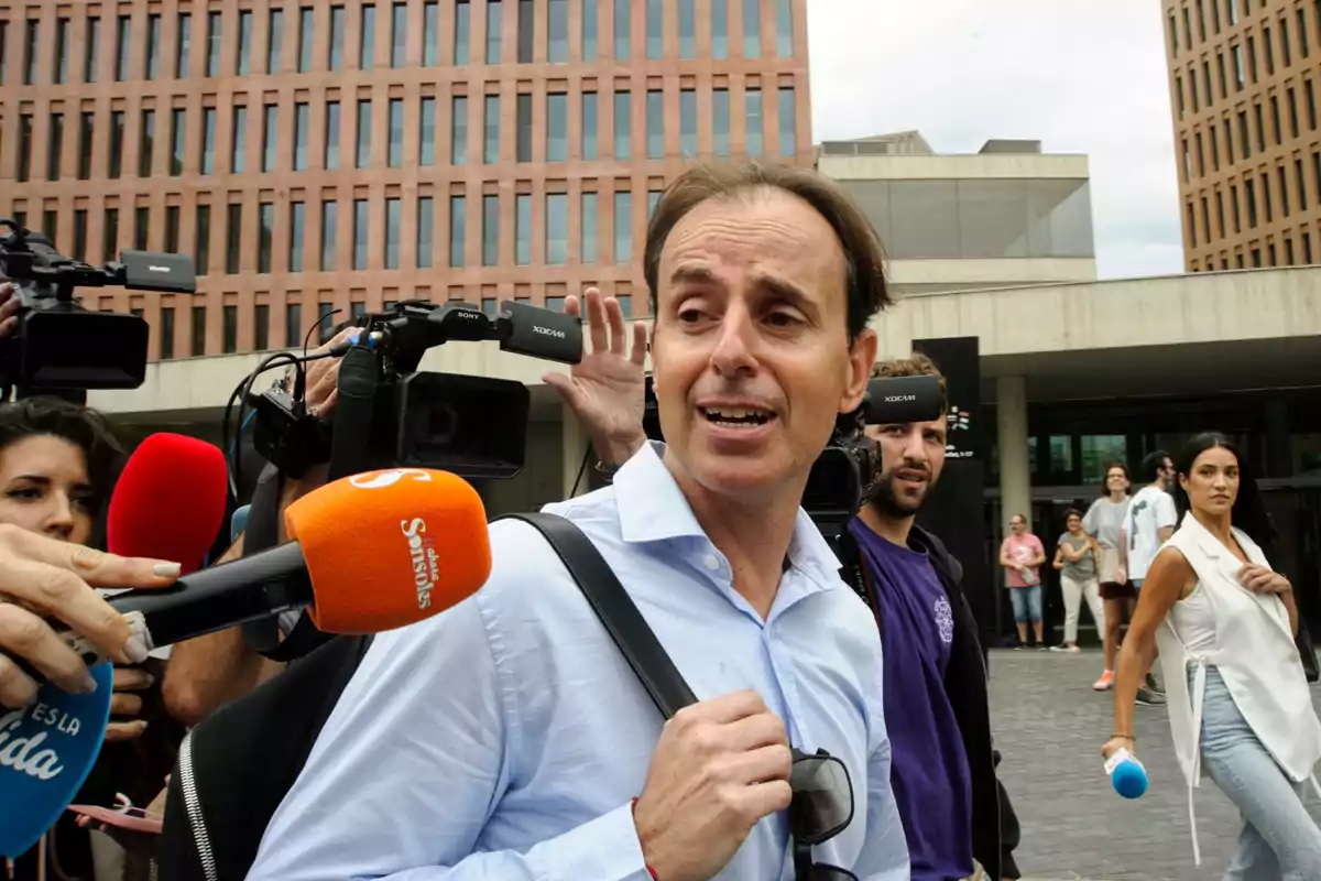 Un hombre con camisa azul claro es entrevistado por varios periodistas con micrófonos y cámaras frente a un edificio de oficinas.