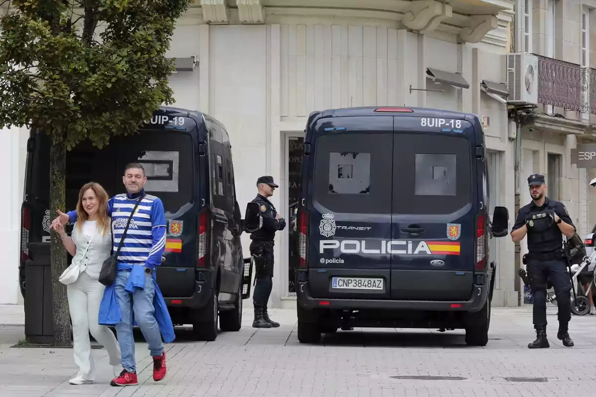 Una pareja camina frente a dos furgonetas de la policía estacionadas en una calle, mientras dos agentes están de pie cerca de los vehículos. Una pareja camina frente a dos furgonetas de la policía estacionadas en una calle, mientras dos agentes están de pie cerca de los vehículos.