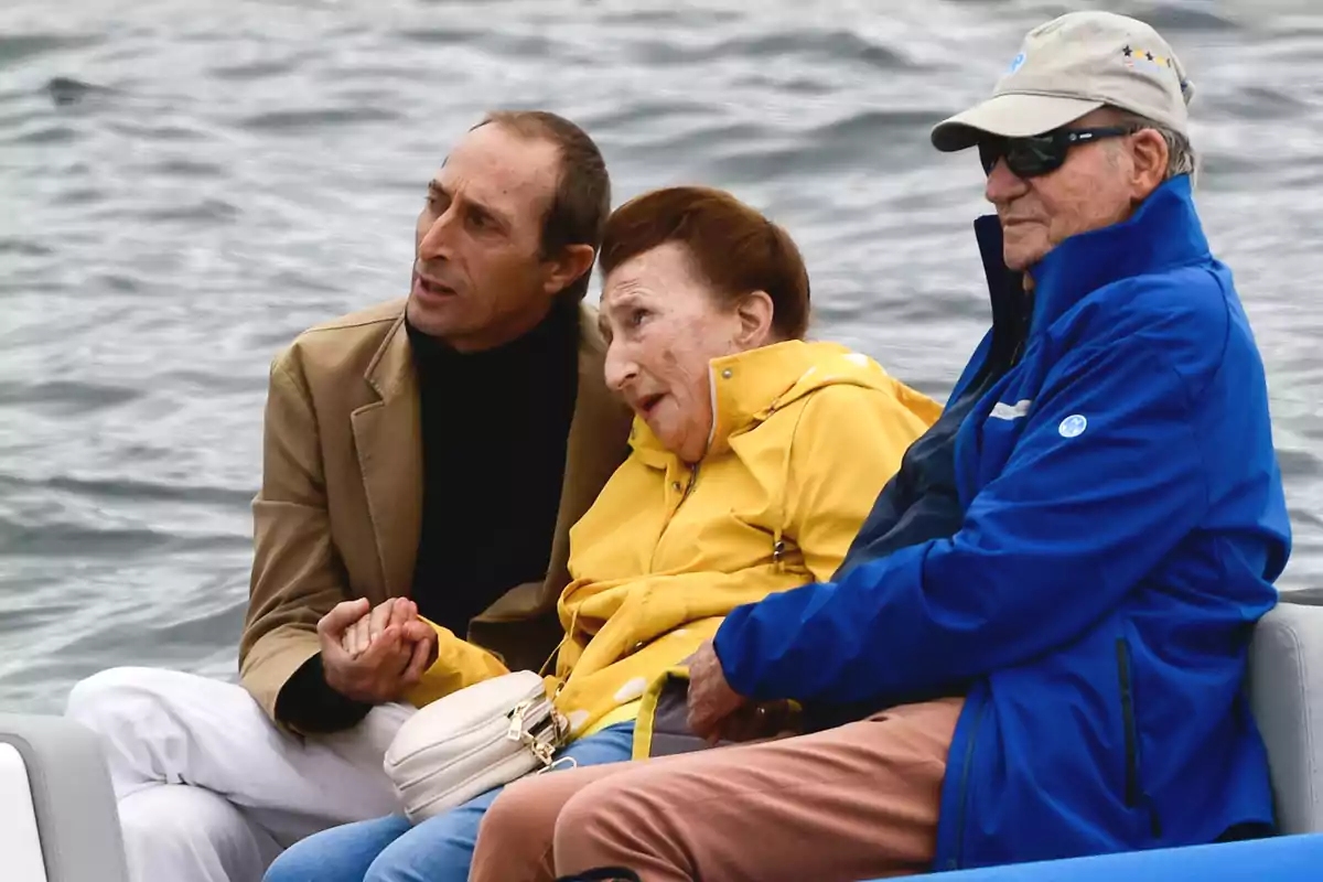 Tres personas sentadas en un bote sobre el agua, una de ellas lleva una chaqueta amarilla, otra una chaqueta azul y gorra, y la tercera un abrigo marrón. Tres personas sentadas en un bote sobre el agua, una de ellas lleva una chaqueta amarilla, otra una chaqueta azul y gorra, y la tercera un abrigo marrón.