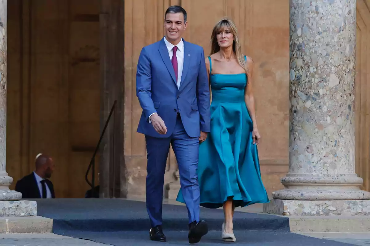 Una pareja elegantemente vestida camina de la mano en un entorno arquitectónico clásico.