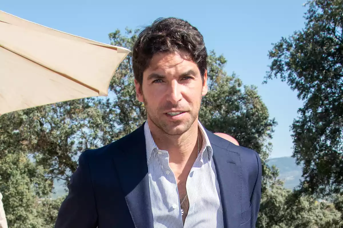 Hombre con traje azul y camisa a rayas en un entorno al aire libre con árboles y cielo despejado.