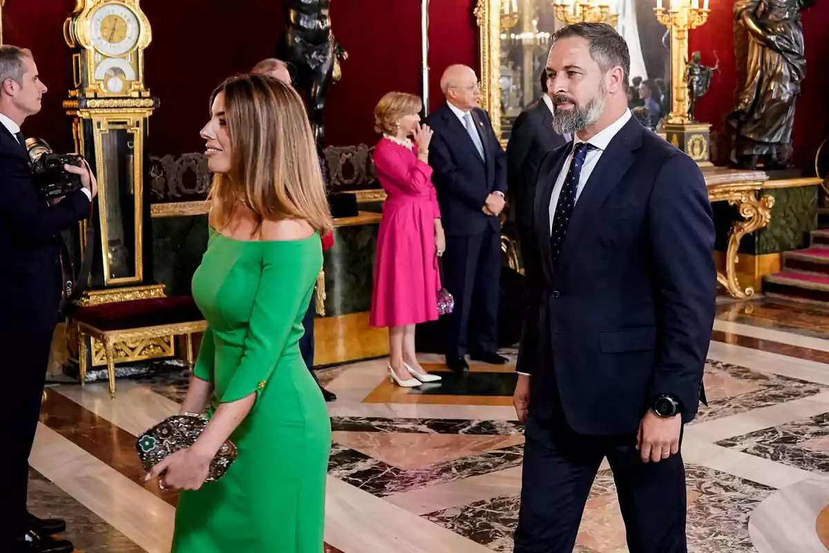 Un grupo de personas elegantemente vestidas se encuentra en un salón decorado con relojes y estatuas doradas. Un grupo de personas elegantemente vestidas se encuentra en un salón decorado con relojes y estatuas doradas.