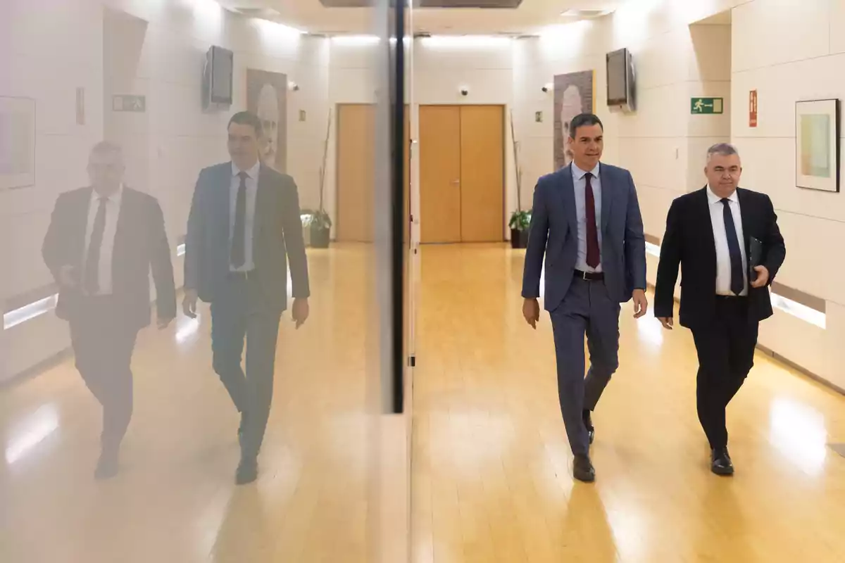 Dos hombres con traje caminan por un pasillo iluminado con suelo de madera y una pared de cristal que refleja su imagen Dos hombres con traje caminan por un pasillo iluminado con suelo de madera y una pared de cristal que refleja su imagen