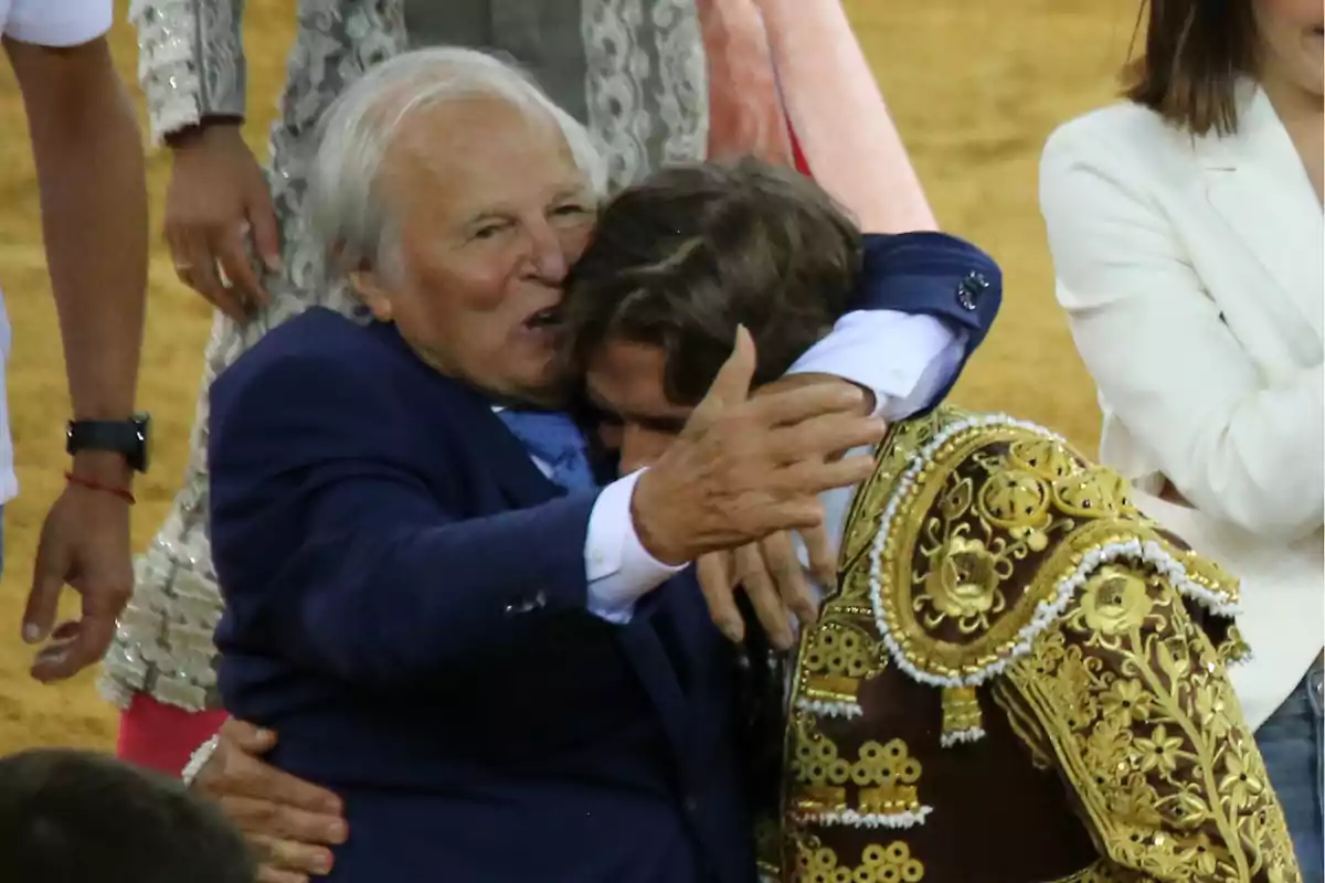 Un hombre mayor abraza a un torero vestido con un traje tradicional en una plaza de toros, rodeados de otras personas. Un hombre mayor abraza a un torero vestido con un traje tradicional en una plaza de toros, rodeados de otras personas.